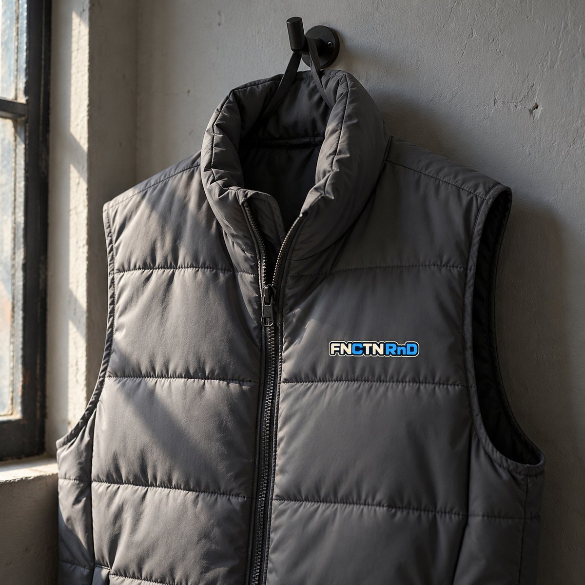 The Puffer Vest in Shadow Grey on a hook by a window